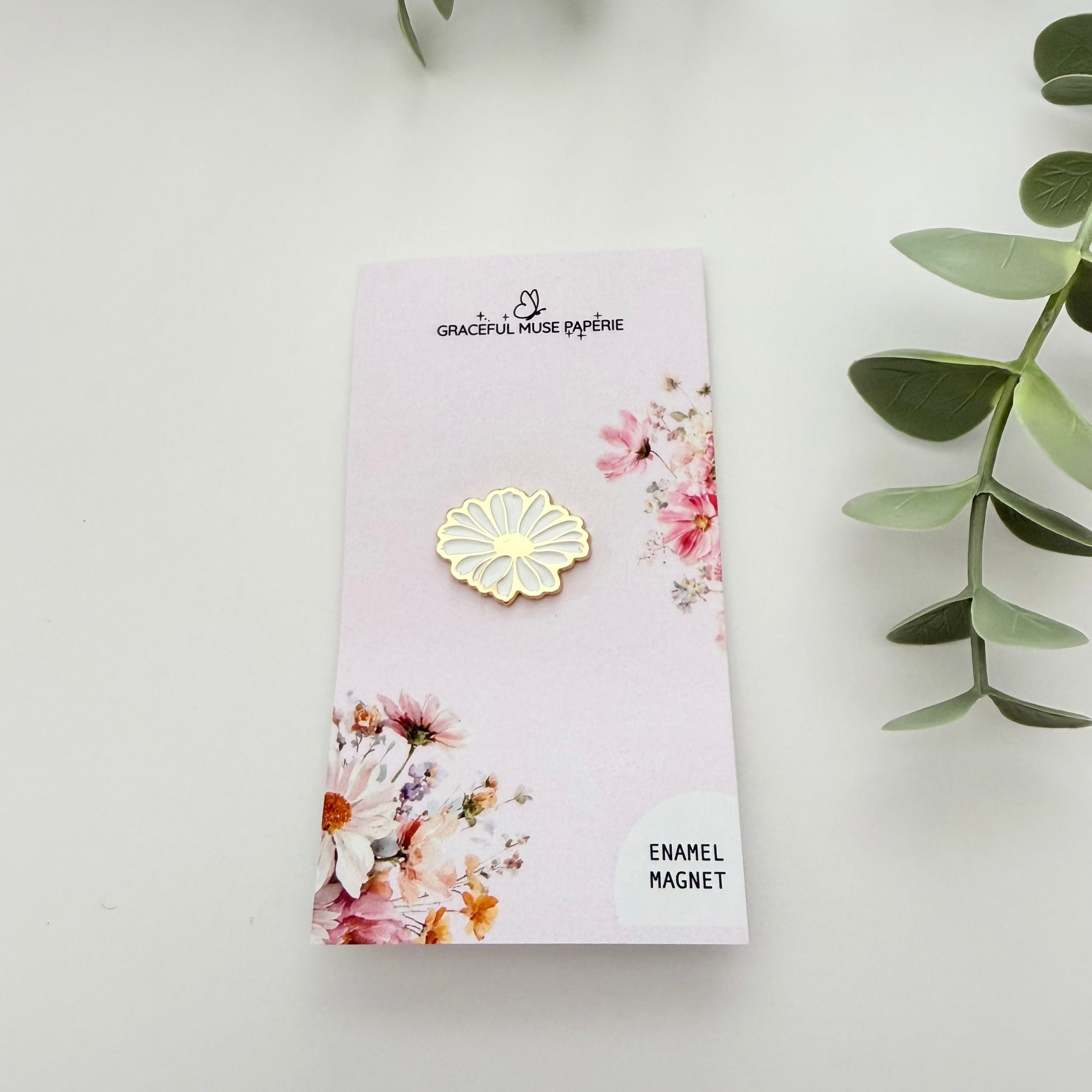 A small gold daisy-shaped enamel magnet displayed on a pink backing card with floral illustrations, photographed on a white surface with green leaves in the corners.