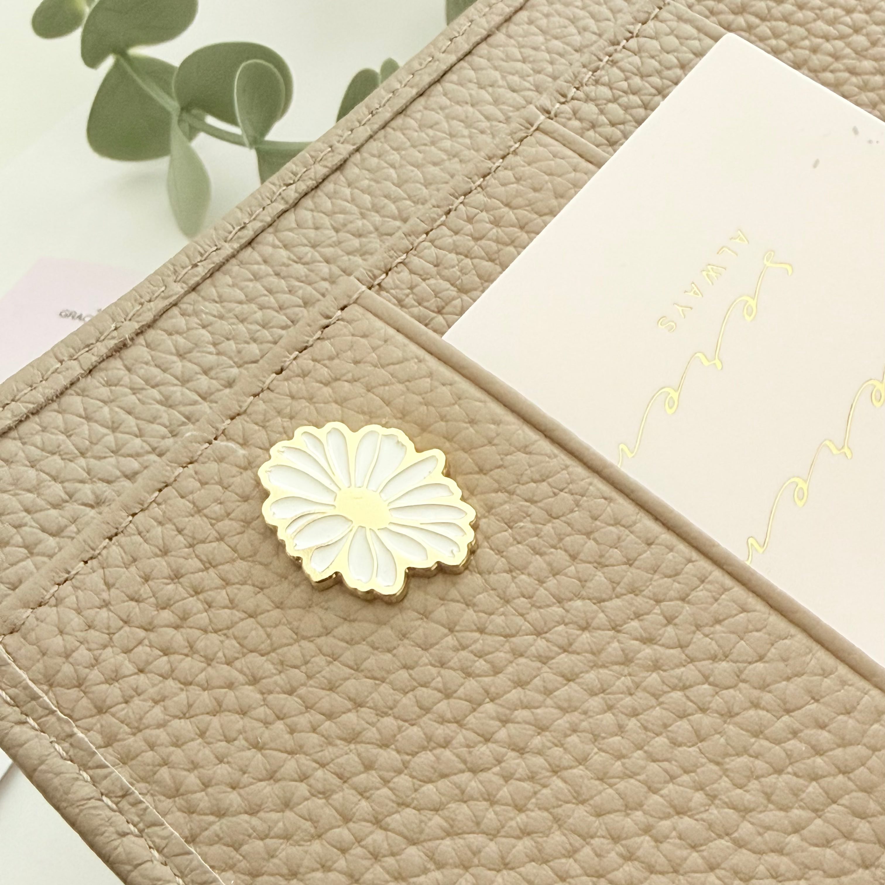A close-up of a beige pebbled leather organizer or pouch with a white flower-shaped metal pin on the front and a cream card slot holding a white card.