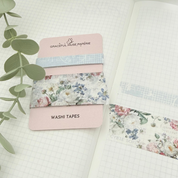 A set of floral and patterned washi tapes displayed on a pink card, styled on an open grid notebook with green eucalyptus leaves.