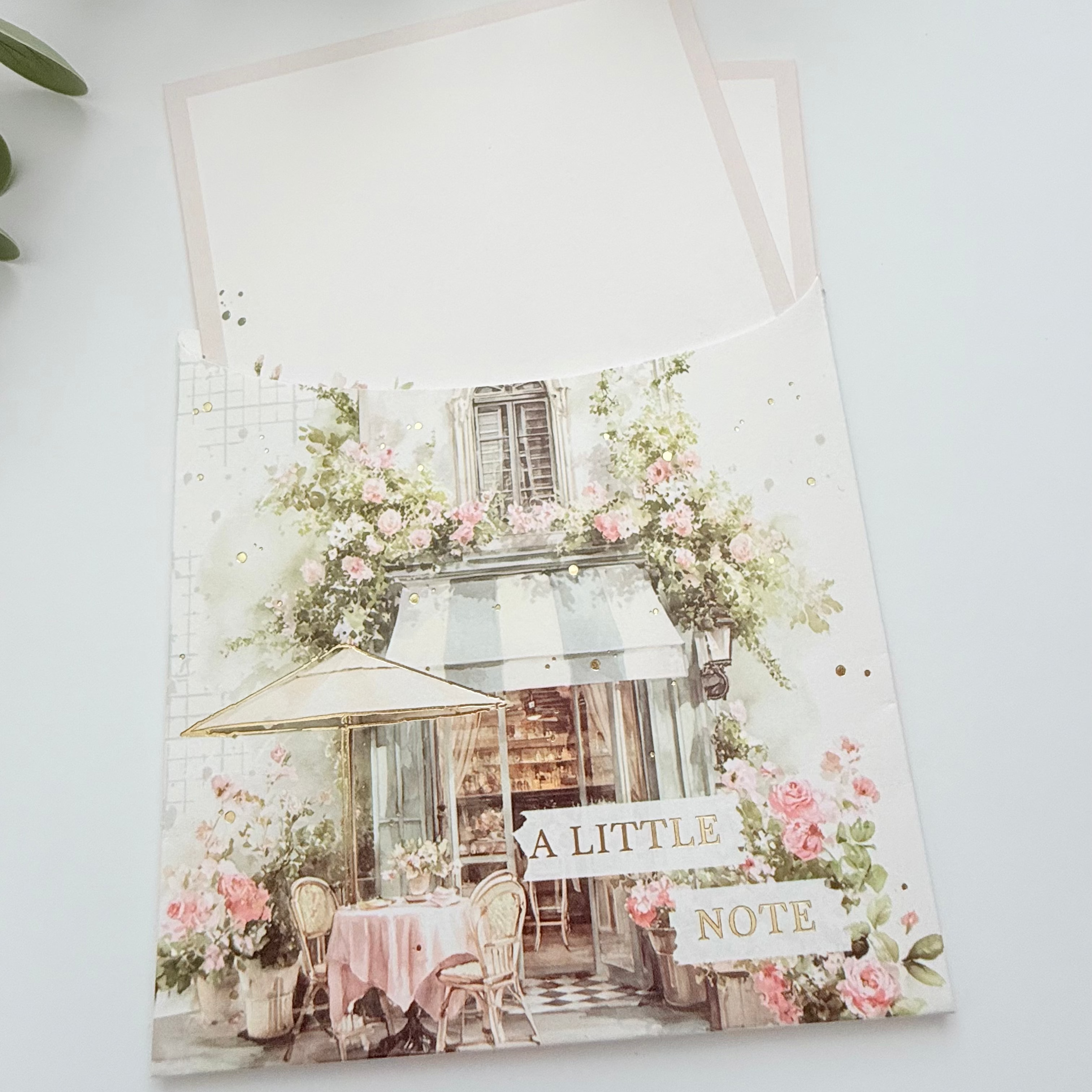 A square greeting card with a French cafe illustration and the words 'A LITTLE NOTE' on the front, shown with a matching blank card partially tucked behind it on a light background with green leaves at the top.