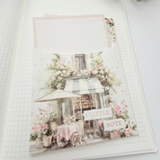 An open grid journal spread showing a decorative cafe-themed page with pink flowers, a small table and chair, and a blank card slot, styled with green leaves at the top.