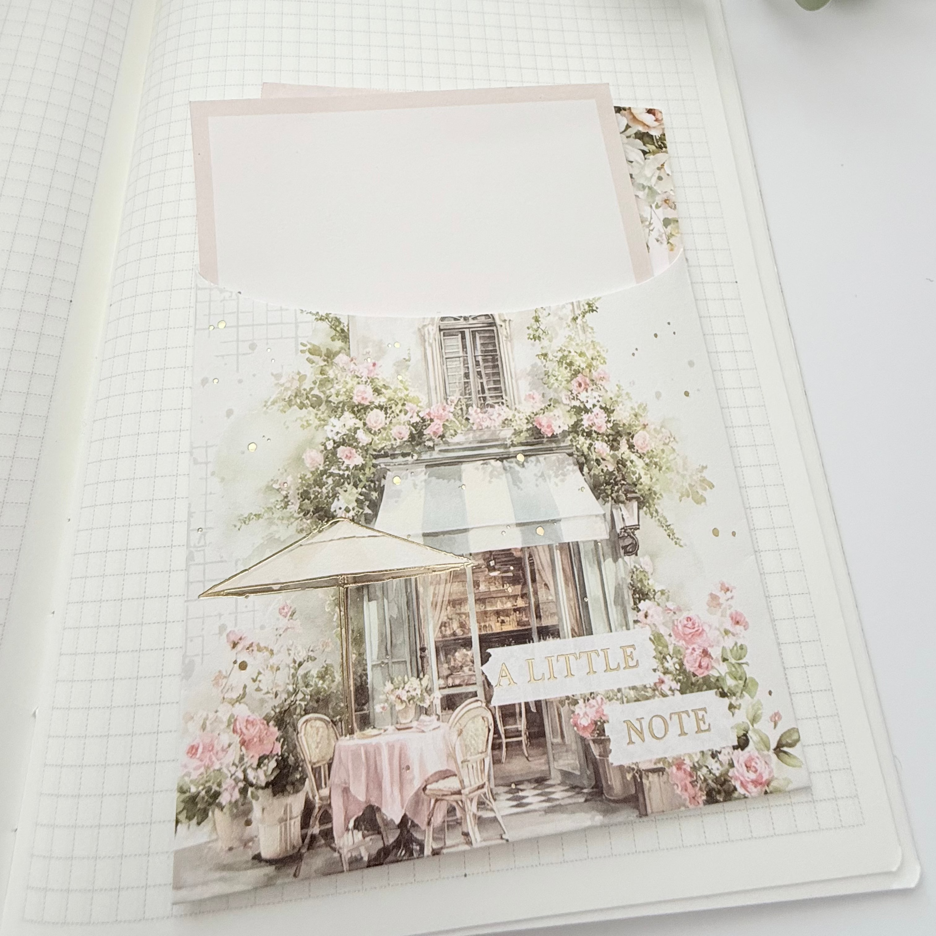 An open grid journal spread showing a decorative cafe-themed page with pink flowers, a small table and chair, and a blank card slot, styled with green leaves at the top.
