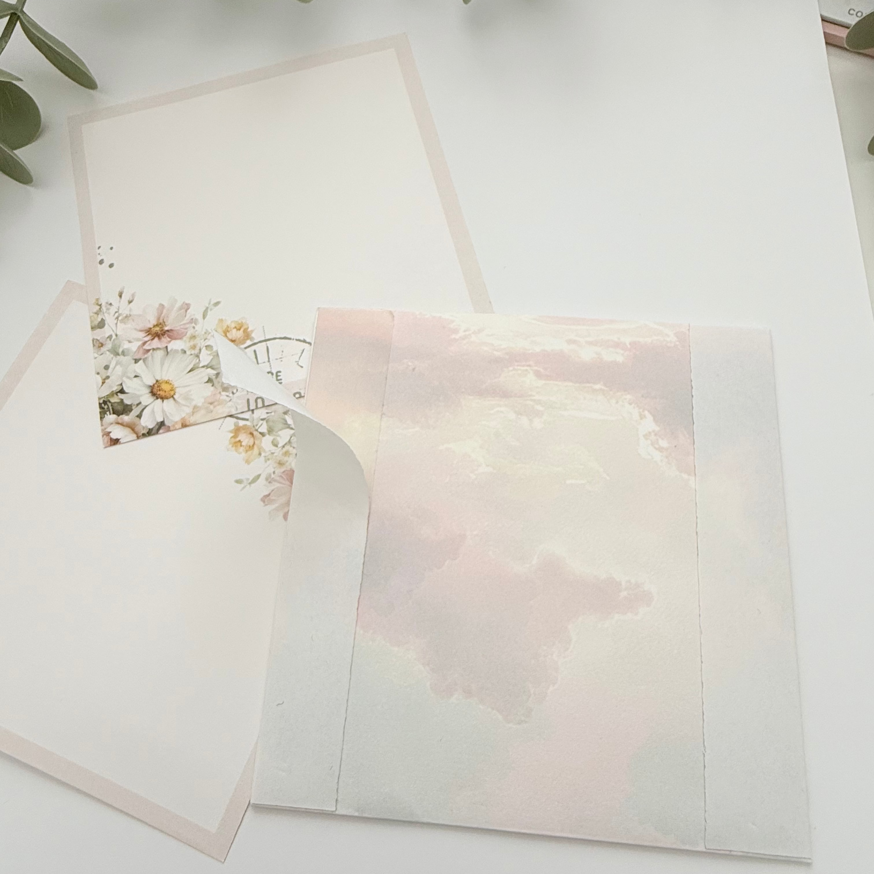 A styled flat-lay of a square greeting card or art print featuring a soft pastel pink and cream cloud design, shown with a matching floral envelope on a white surface with green eucalyptus leaves.